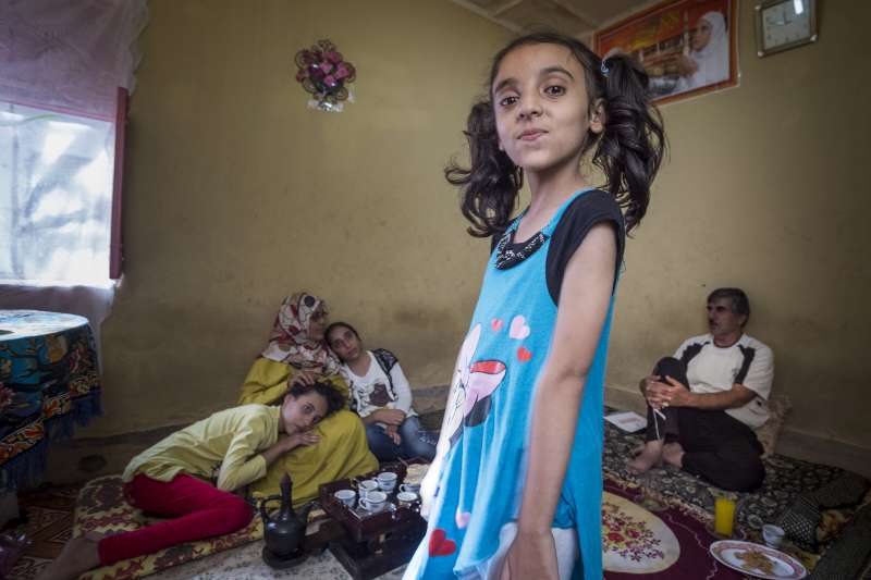 Nora (eight years old, in blue dress) is the youngest of the three daughters of Husam and Nemah. In the background, sitting on a mattress, left to right: daughter Kholod (11 years old), mother Nemah and Asmaa (14 years old). Father Husam is sitting on the right.
