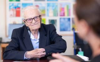 Harry Leslie Smith sits at table in front of a colourful wall of pictures