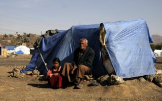 An older man sits outside of a tent covered by a blue tarp in a refugee camp in Yemen and to his left a young girl is crouched wearing a red dress