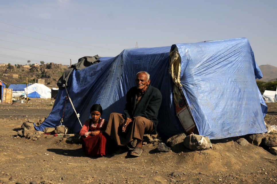 An older man sits outside of a tent covered by a blue tarp in a refugee camp in Yemen and to his left a young girl is crouched wearing a red dress