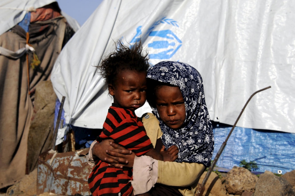 a refugee child from Yemen carrying her sibling standing in front of temporary shelter tents with U.N.H.C.R. branding on it