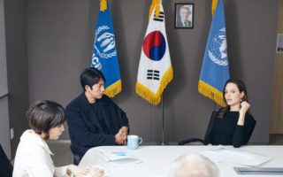 Special envoy Angelina Jolie and goodwill ambassador Jung Wuo-Sung sitting at a table in front of three flags, the two outer flags are UN flags and the middle flag is the Korean Flag. There is a woman sitting to the left of them also at the table.