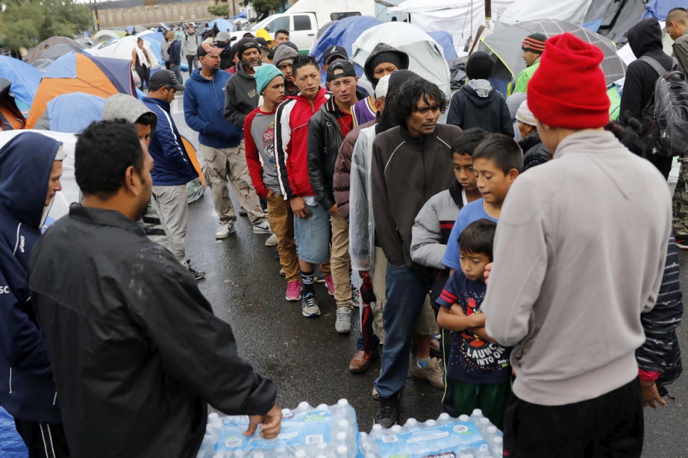 group of asylum seekers lining up amidst a village of tents to receive water bottles from three men at the front of the line as new asylum protocols announced