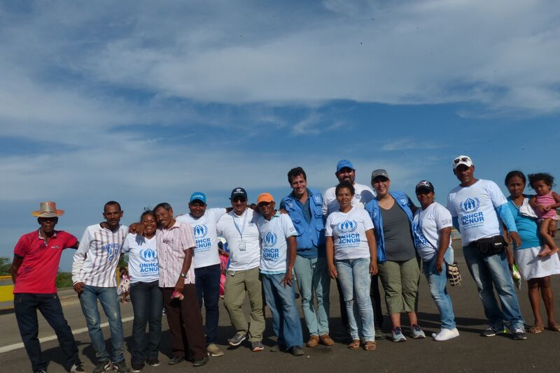 A group of people are standing against a blue sky posing happily with many wearing UNHCR branded tshirts and blue vests and hats