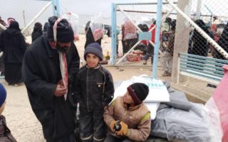 A man standing beside two children, one child stands the other one sits, behind them is a scene of a refugee resettlement camp.