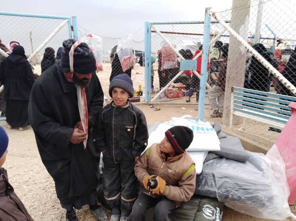 A man standing beside two children, one child stands the other one sits, behind them is a scene of a refugee resettlement camp.