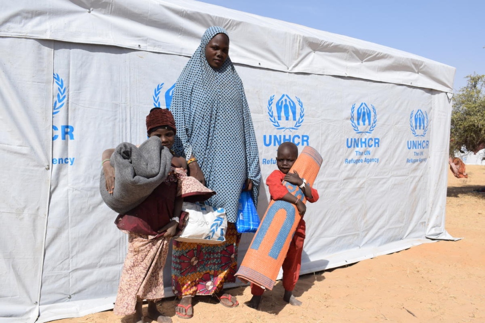 A woman standing in front of a tent with the UNHCR logo on it holding on to her two children one on each side who are holding blankets and mats