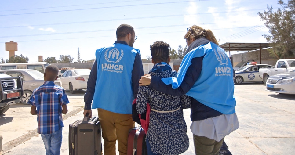 two children and two adults wearing blue vests with UNHCR logo emblazoned on the back walk away from the camera, the children have luggage and the adults are helping them carry the luggage as they walk across an outdoor parking lot