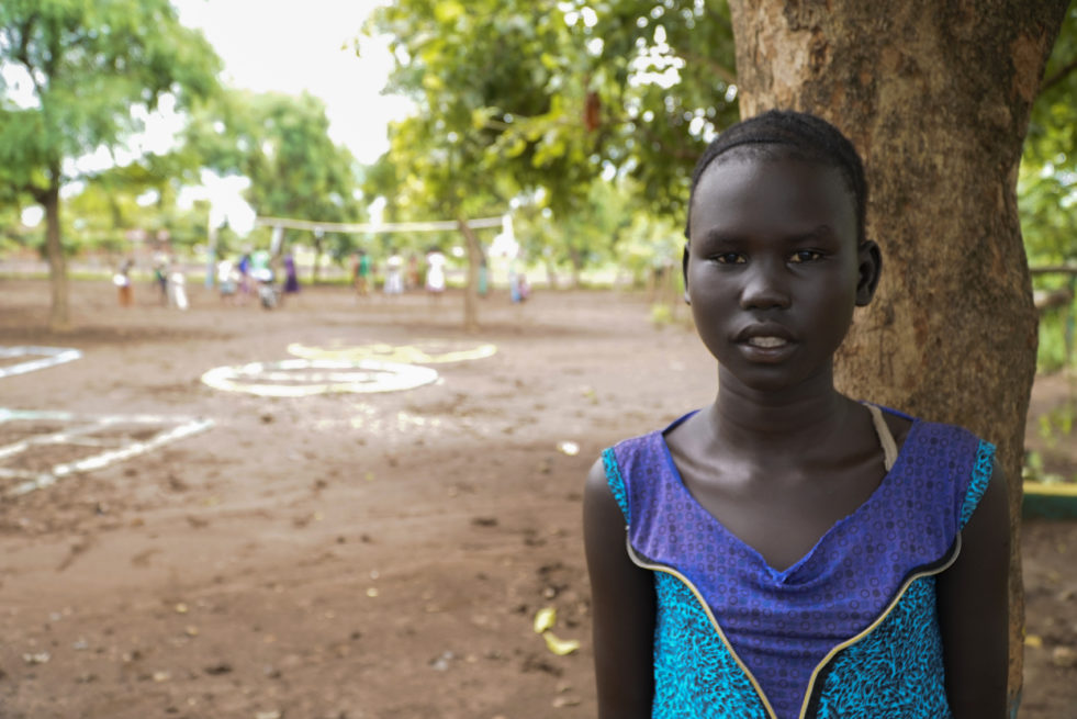 Young South Sudanese refugee girl poses in front of a camera in a blue and purple dress against the backdrop of an open field surrounded by trees
