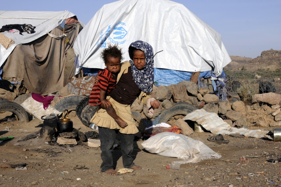a young girl carries a baby on her hip, both of them are dirty and disheveled and are standing barefoot on a dirt road in front of a makeshift tent that has UNHCR branding on it.