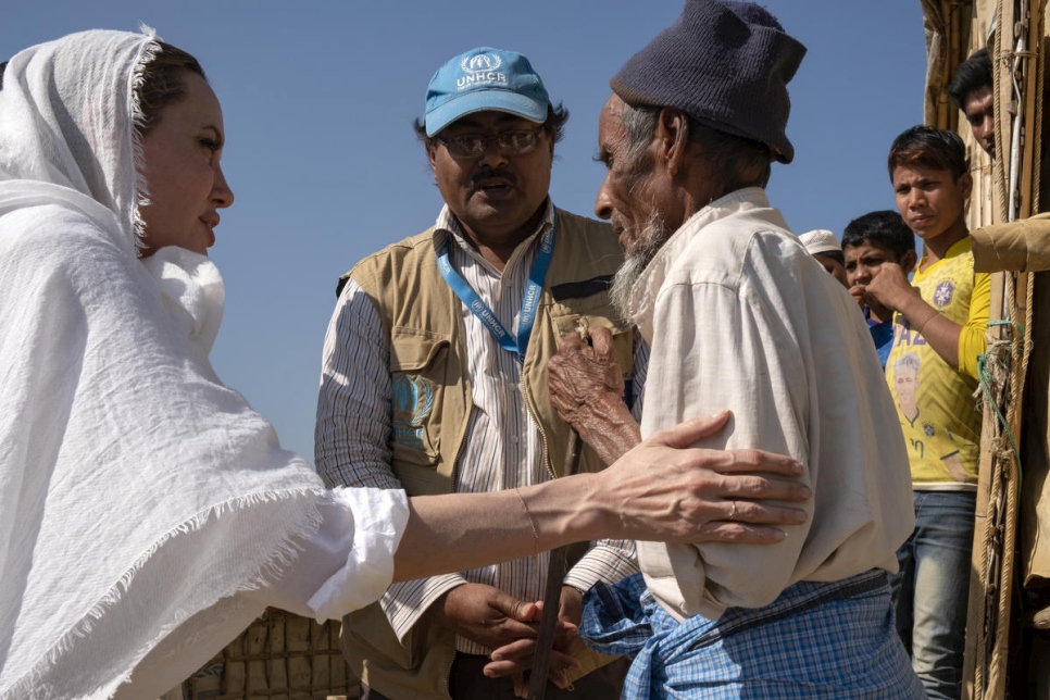 Woman in head scarf touches man's elbow in gesture of support while a man stands between them wearing UNHCR branded clothing and watches the moment unfold.