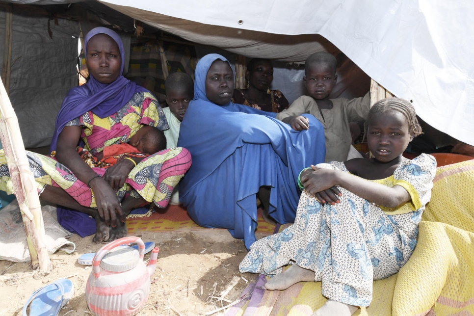 two women sitting underneath a makeshift tent, one is nursing a baby the other is surrounded by three young children. There is another woman who is sitting in the very back of the tent.