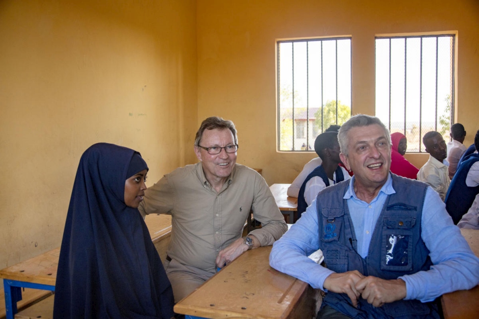 man in blue shirt and vest smiles looking at something behind a camera while he sits at a school desk with a woman in a black hijab and a man wearing a brown shirt, glasses and khaki pants