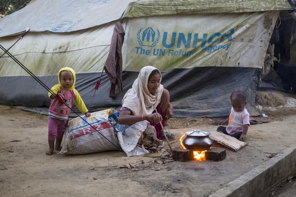 a woman sits on the ground watching a small silver pot cook on a small fire . she is outside and she is sitting on a sack with two children on either side of her. behind her is a tent branded with UNHCR