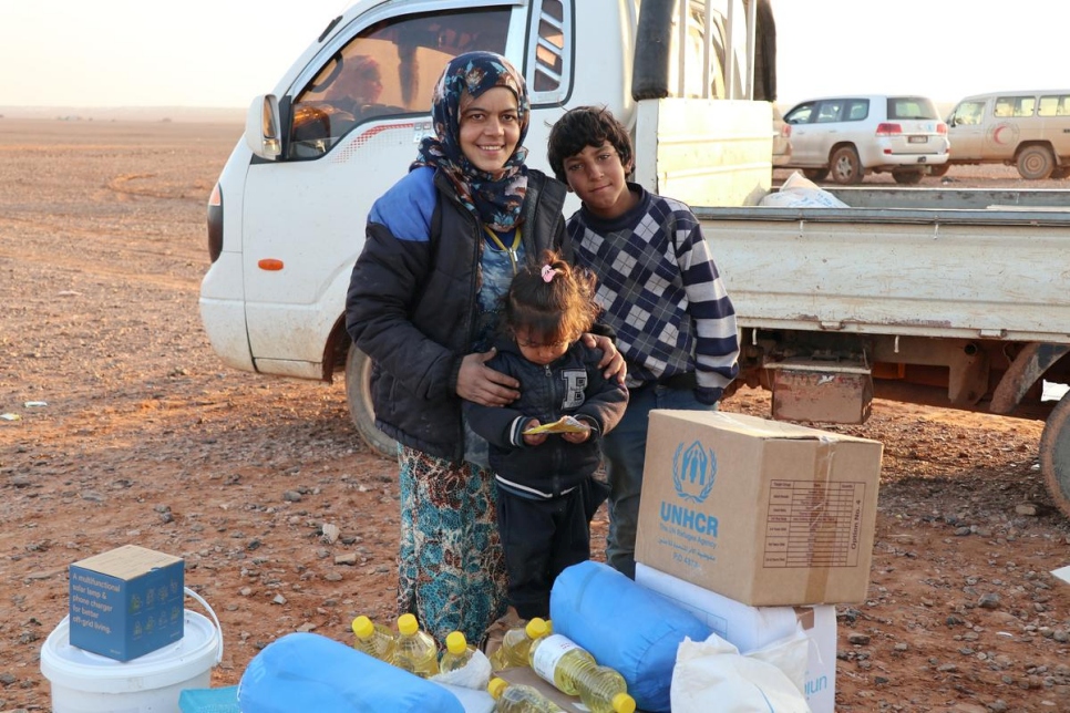 A mother and her two children stand infront of a white truck in a desert surrounded by supplies that have UNHCR branding on them