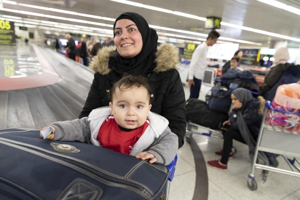 a woman wearing a black hijab pushes a baggage cart with her baby sitting in the cart, they are beside a luggage carousel at an airport
