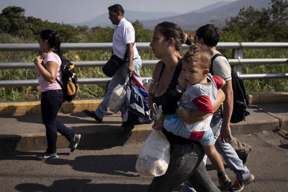 a group of people walk on what appears to be a bridge, closest to the camera is a woman carrying a plastic bag full of supplies and a child on her hip, she is wearing grey denim pants and a black tank top, her hair is tied back in a tight bun