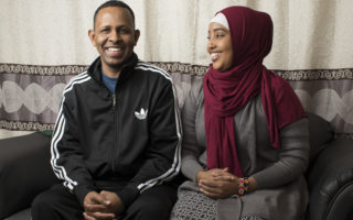 A man and a woman sit on a black leather couch with happy expressions on their faces