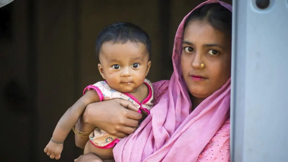 A young mother wearing a pink headscarf and a gold nose piercing holds a baby in her arms