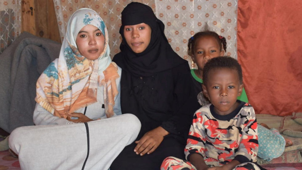 Two women wearing hijabs sit on a bed beside each other with two children sitting to their left