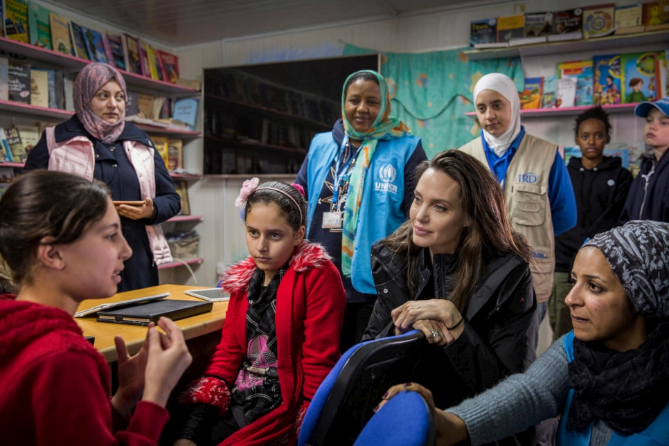 A woman listens intently to a young girl, beside her is another young girl wearing a red jacket with a flower in her hair, she is also listening intently, on the other side is a woman wearing a black and white head scarf and turquoise ribbed sweater behind them is a group of five people with one of them wearing a blue UNHCR vest 