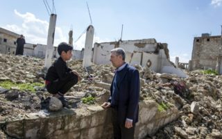 A man speaks to a young boy perched on the ledge of a crumbling wall, behind them is the remnants and rubble of a war-torn community