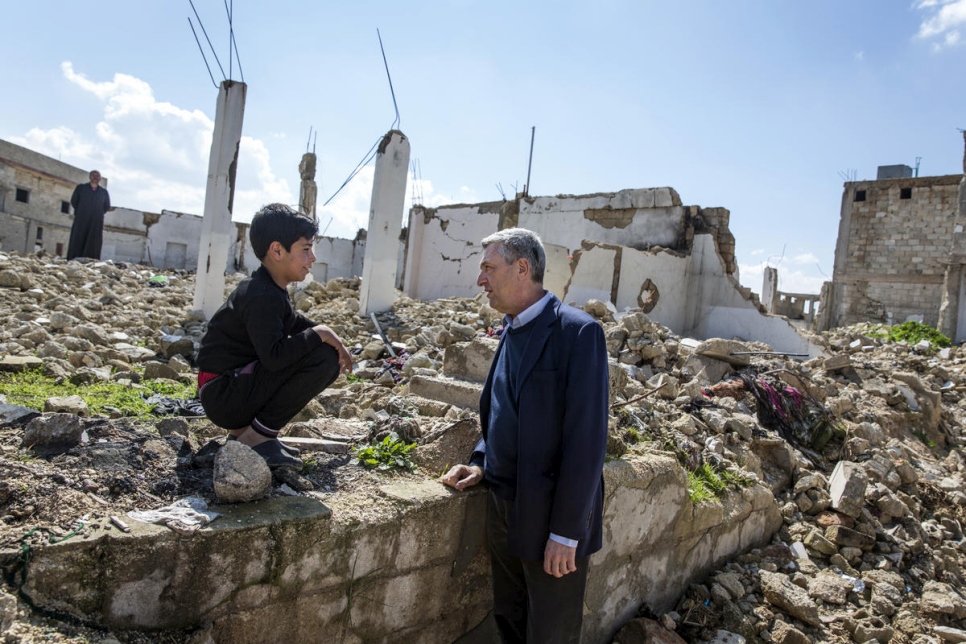 A man speaks to a young boy perched on the ledge of a crumbling wall, behind them is the remnants and rubble of a war-torn community 
