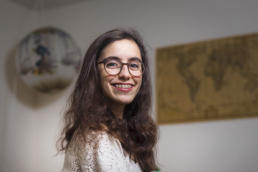 A portrait of a young woman with thick framed glasses, brown long hair, and braces.