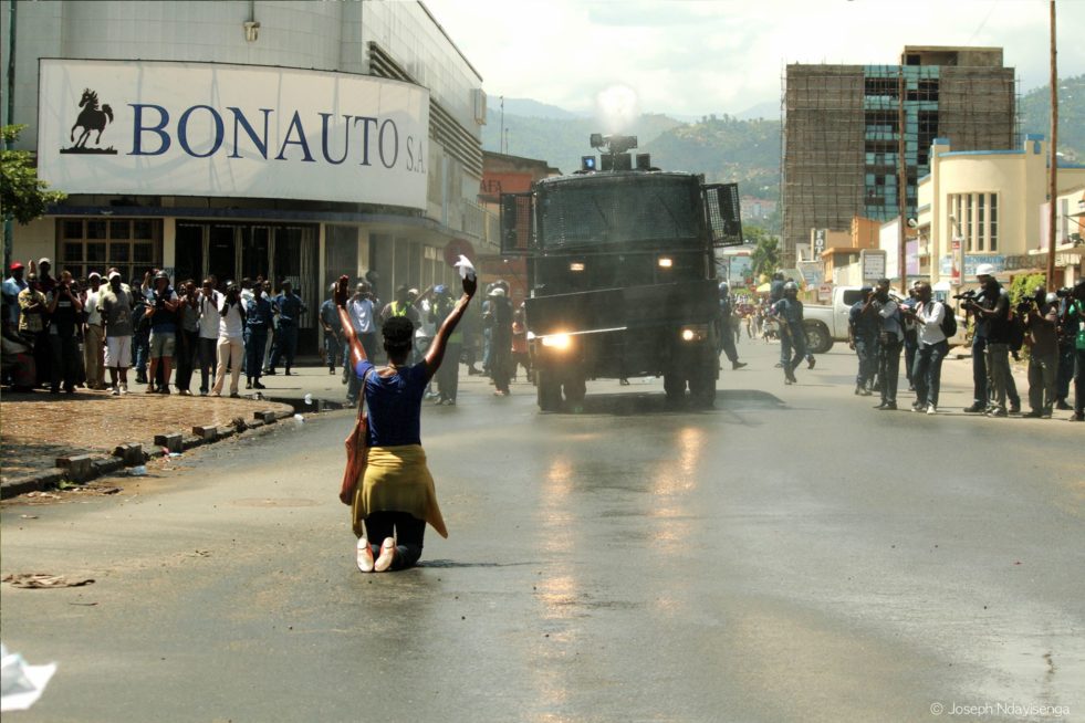 a woman kneels on the street with her back facing the camera with her hands up as if in surrender and an armoured vehicle is on the road heading in her direction, around the vehicle are crowds of people