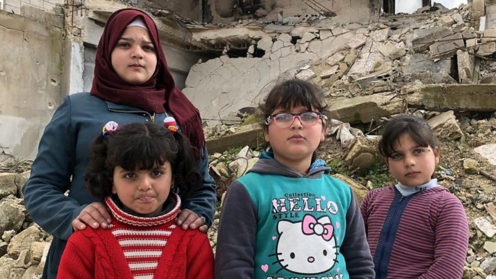 Four young girls stand in front of the rubble and ruined remains of a former building