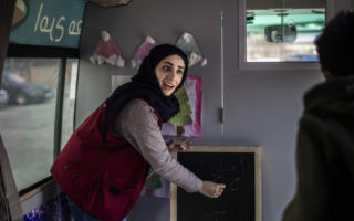 A woman in a black hijab and a maroon vest writes on a small chalkboard as if teaching a classroom