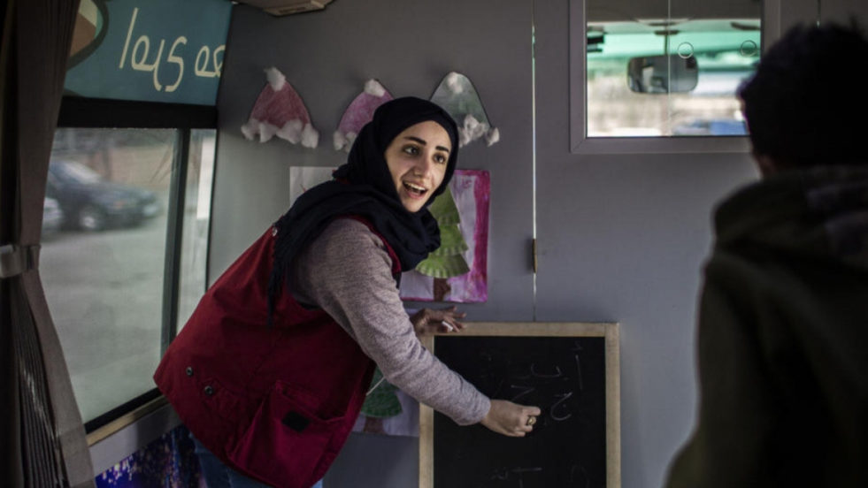 A woman in a black hijab and a maroon vest writes on a small chalkboard as if teaching a classroom