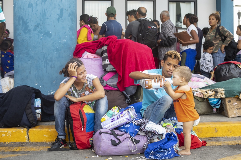 A woman and man sits on the curb of the street while feeding a toddler milk from a carton, behind them is a crowd of people lined up in front of a white wall with windows and stacks of luggage are piled all around