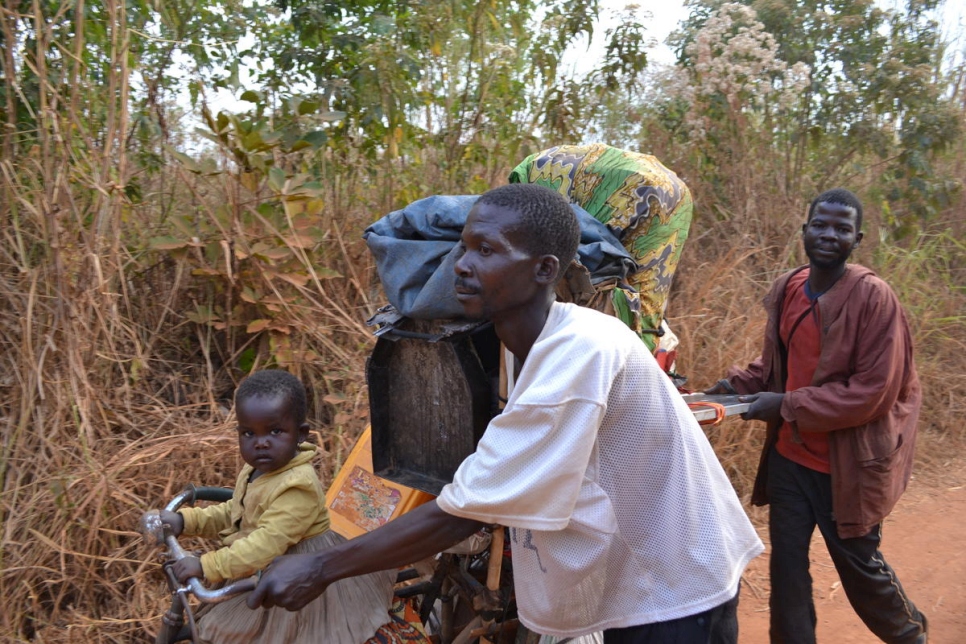 A two men push a wagon stacked with belongings as a baby sits at the front of the wagon, behind them are dried grass and trees, they walk on a dirt road in the DRC