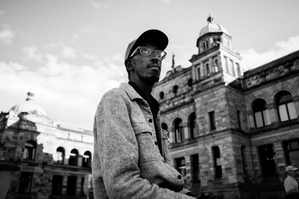 a sitting man wearing glasses and a ball cap looks pensively into the distant horizon. behind him is the BC Legislative Assembly building and the photo is black and white