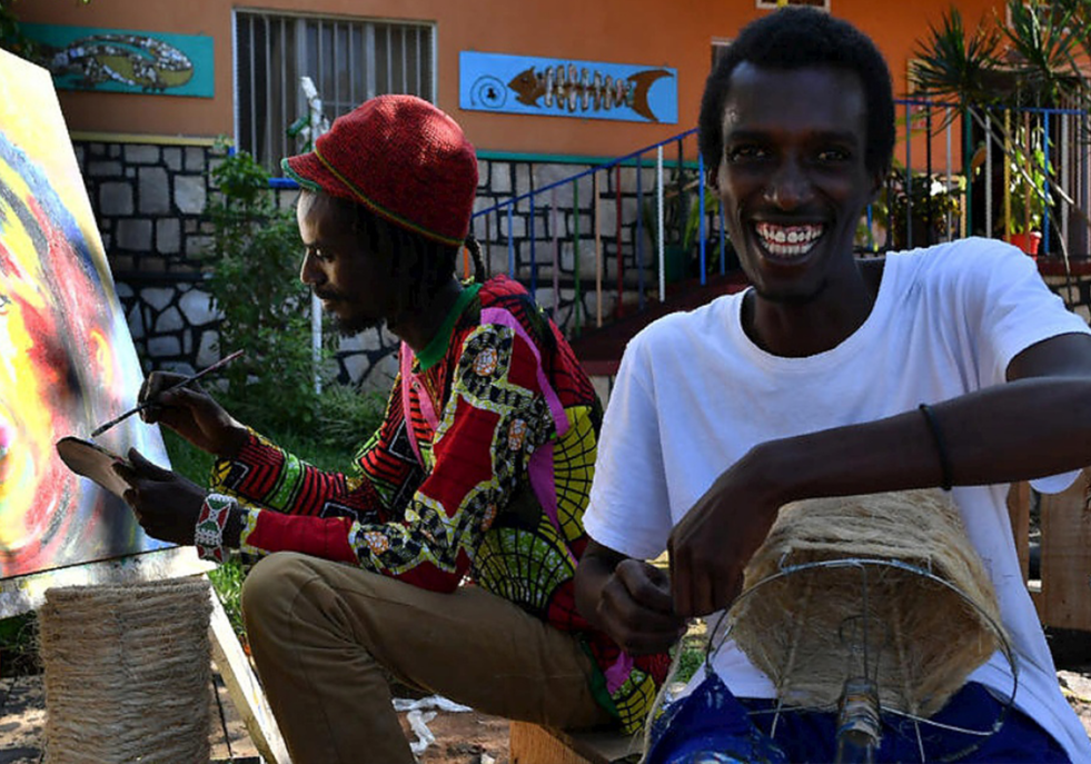 Refugee artists sit be side laughing one painting the other making a lampshade