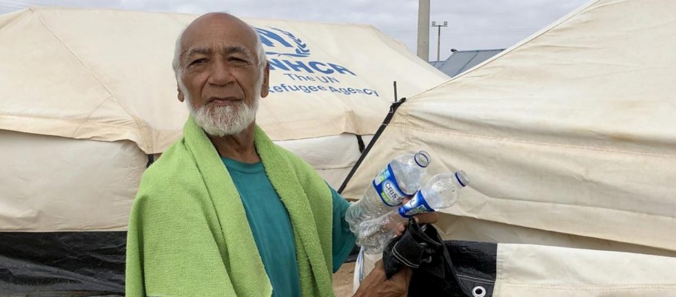 Older venezuelan man with grey beard stands in front of UNHCR branded tends holding two plastic water bottles and a green towel wrapped around his shoulders.