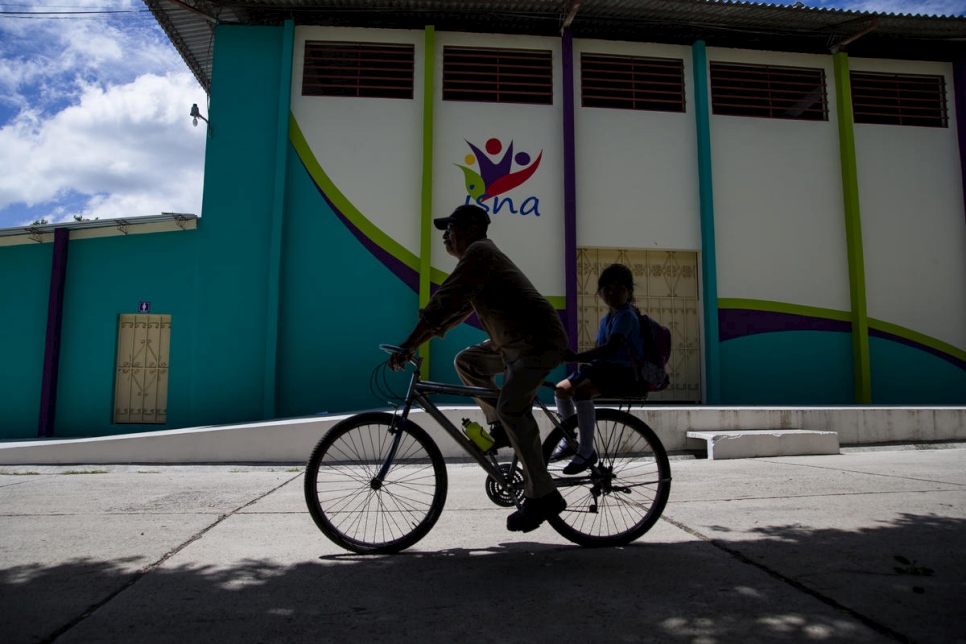 A silhouetted man rides his bike with his daughter seated in a child's seat behind him along the streets of El Salvador