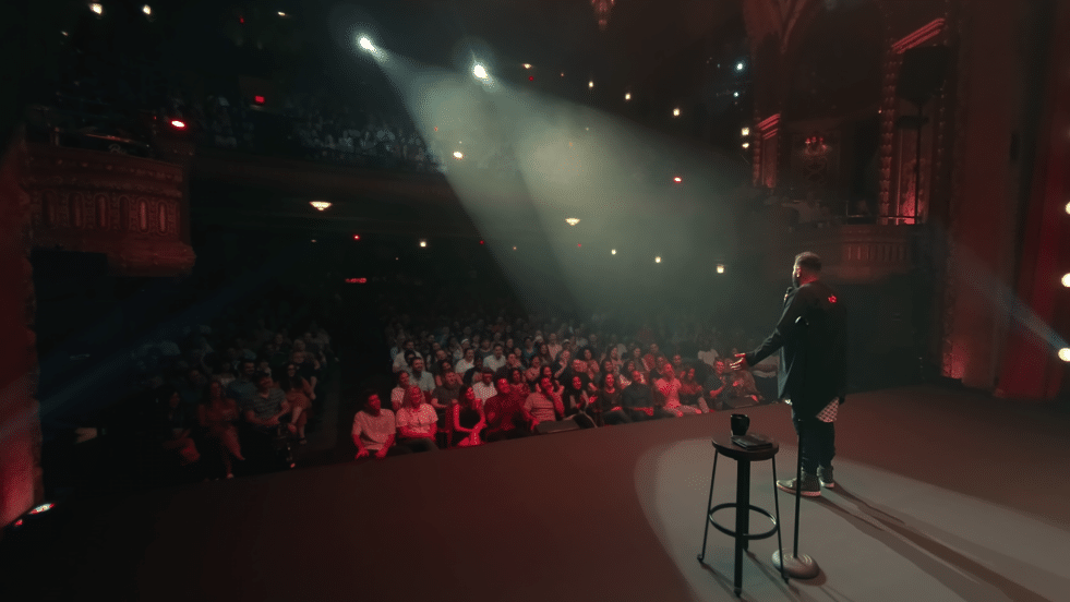 Mo Amer stands on stage looking out at a full theatre