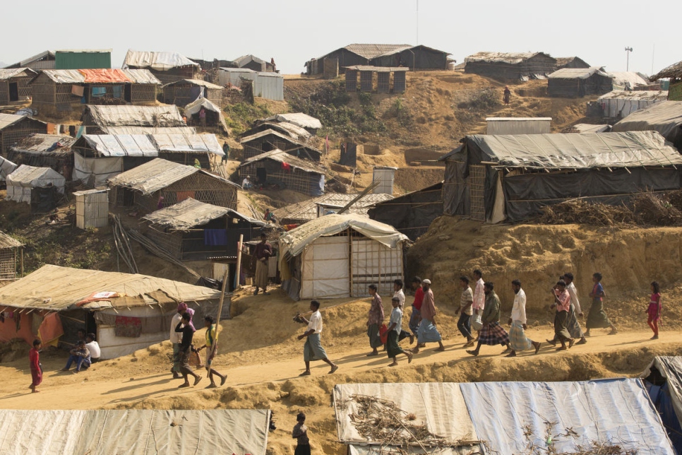 A group of Rohingya refugees walk down a dirt road against the backdrop of Kutupalong