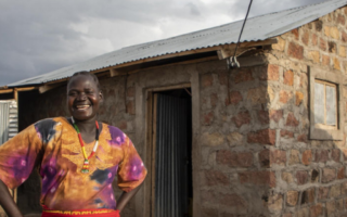 A smiling woman from Kenya stands outside of her house