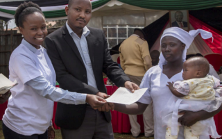 A Kenyan man and woman hand a birth certificate over to a woman carrying a child