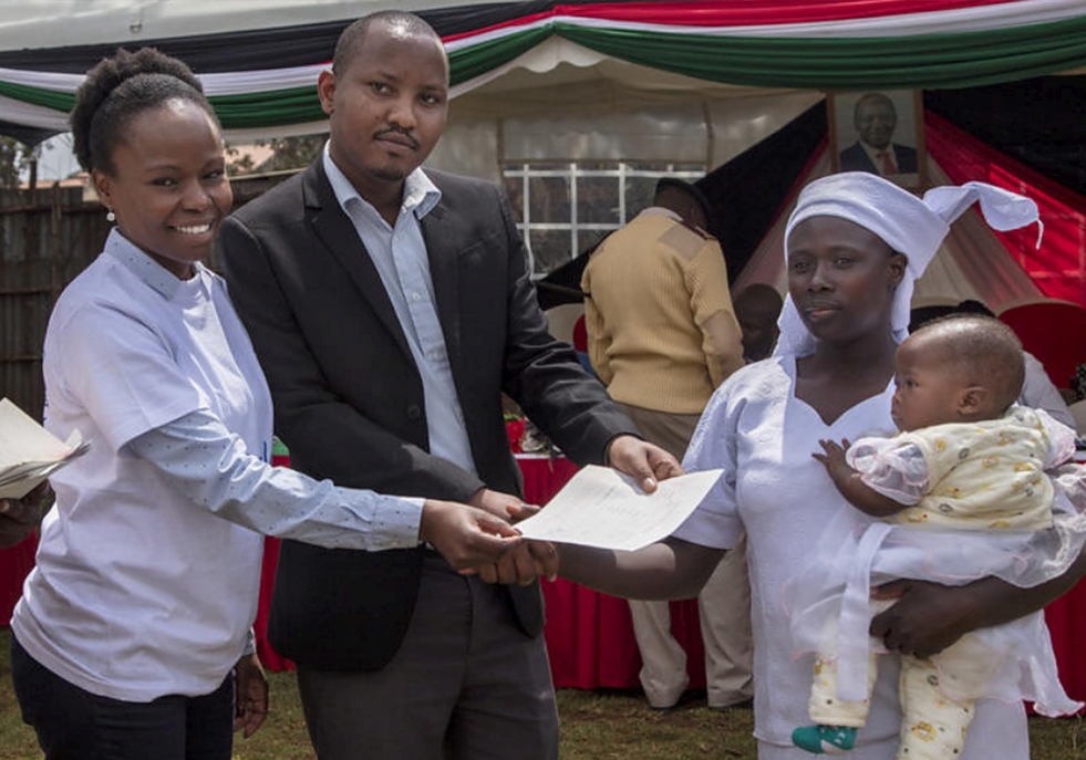 A Kenyan man and woman hand a birth certificate over to a woman carrying a child