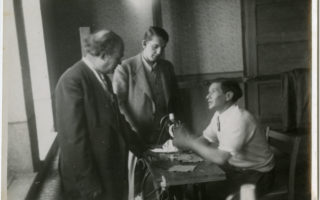 A black and white photo of a tailor sitting at a sewing machine speaking to two men