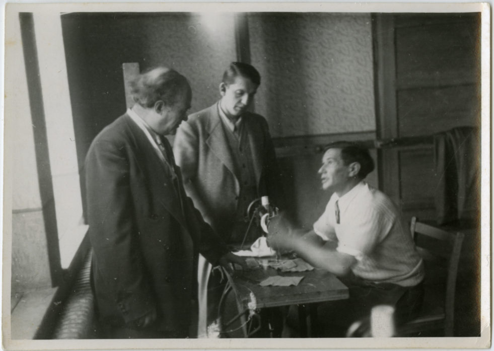 A black and white photo of a tailor sitting at a sewing machine speaking to two men