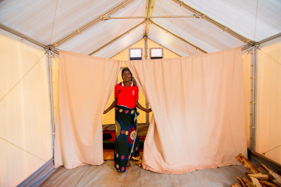 A woman stands in her RHU, Refugee Housing Unit, and poses at a curtain partition