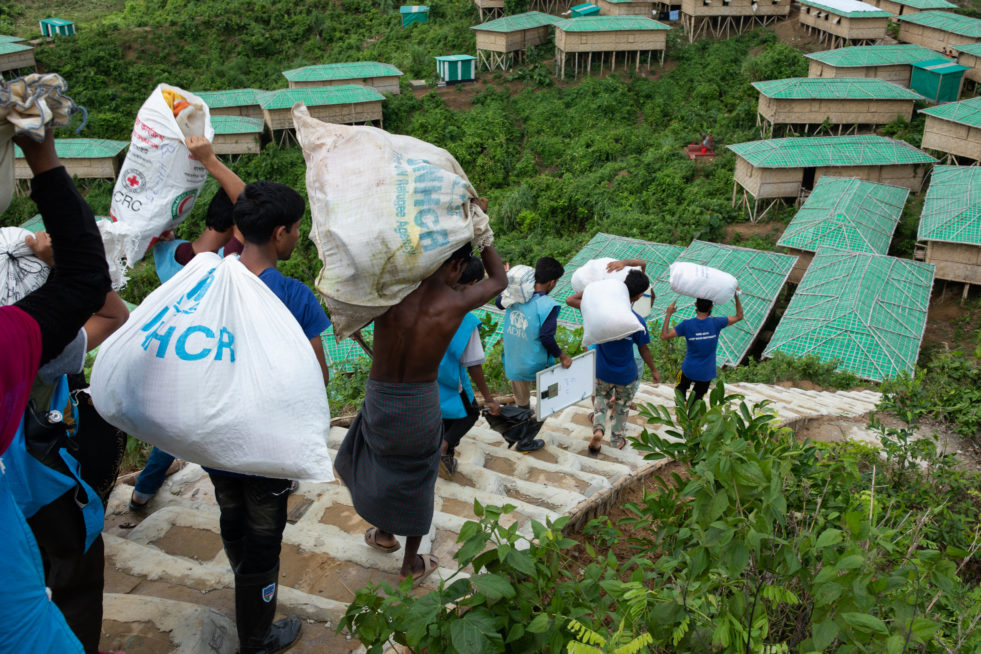 Rohingya refugees carrying sacks with UNHCR branding down a long set of outdoor, dirt stairs