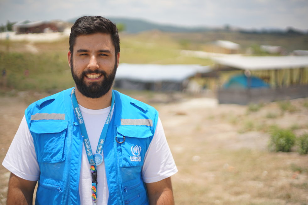 A bearded man stands in front of a building smiling while wearing a UNHCR branded blue vest