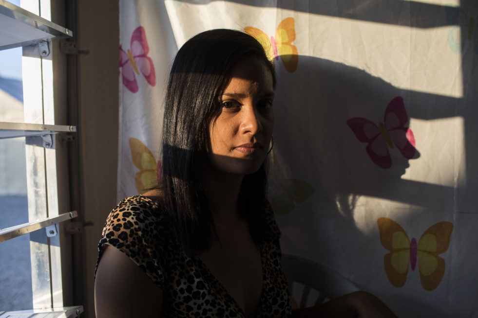 A younh Venezuelan woman stands by a window casting a shadow against a wall of painted butterflies