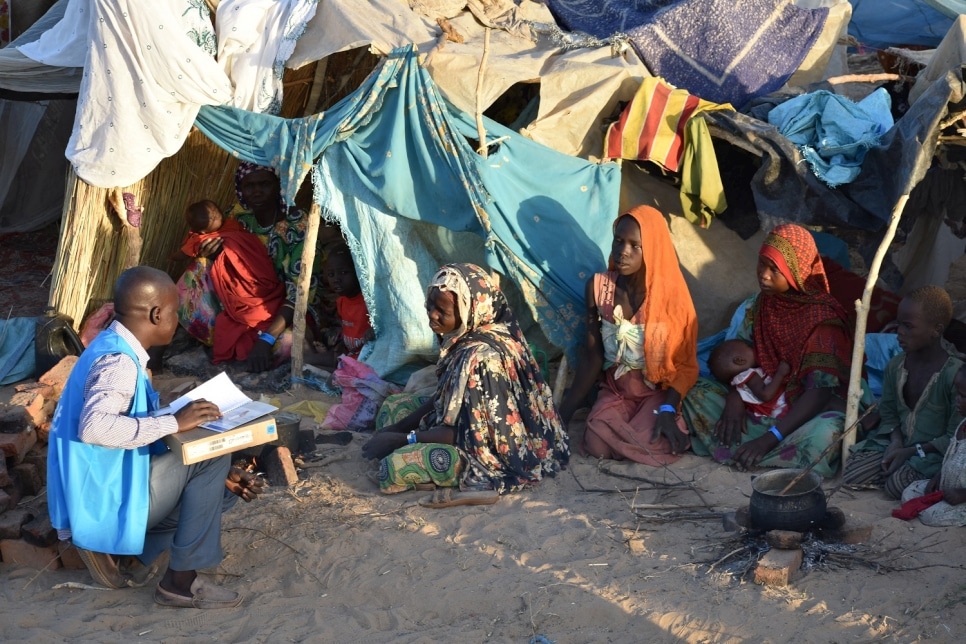 A humanitarian worker in a blue vest speaks to people living in tents from Sudan